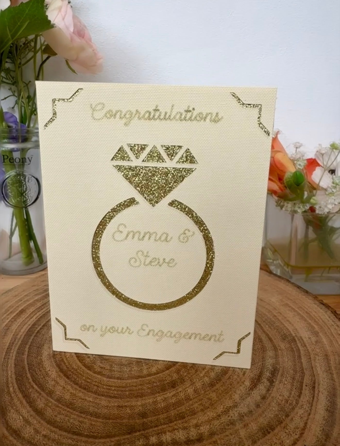 Personalised engagement card with glitter cut-out engagement ring design on cream card. The ring displays the names Emma & Steve inside. Card shown upright on a wooden base with flowers in the background.