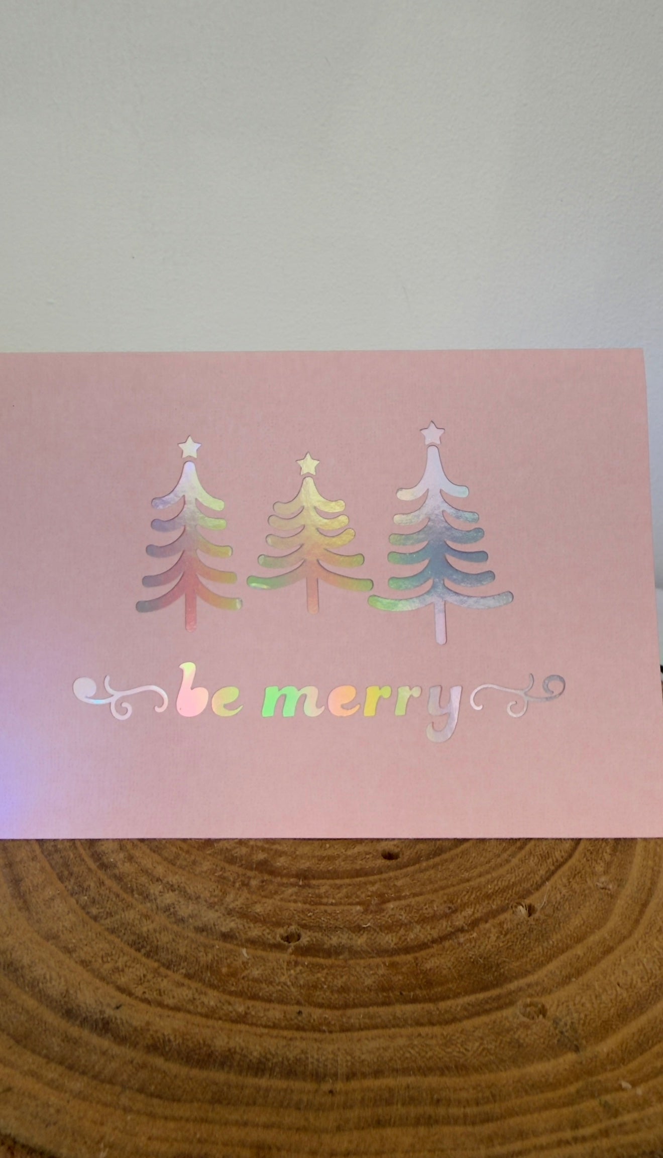 Colorful Christmas tree designs on a pink card with 'be merry' text, placed on a wooden surface.