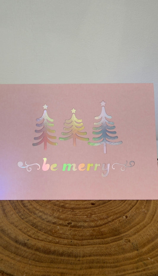 Colorful Christmas tree designs on a pink card with 'be merry' text, placed on a wooden surface.