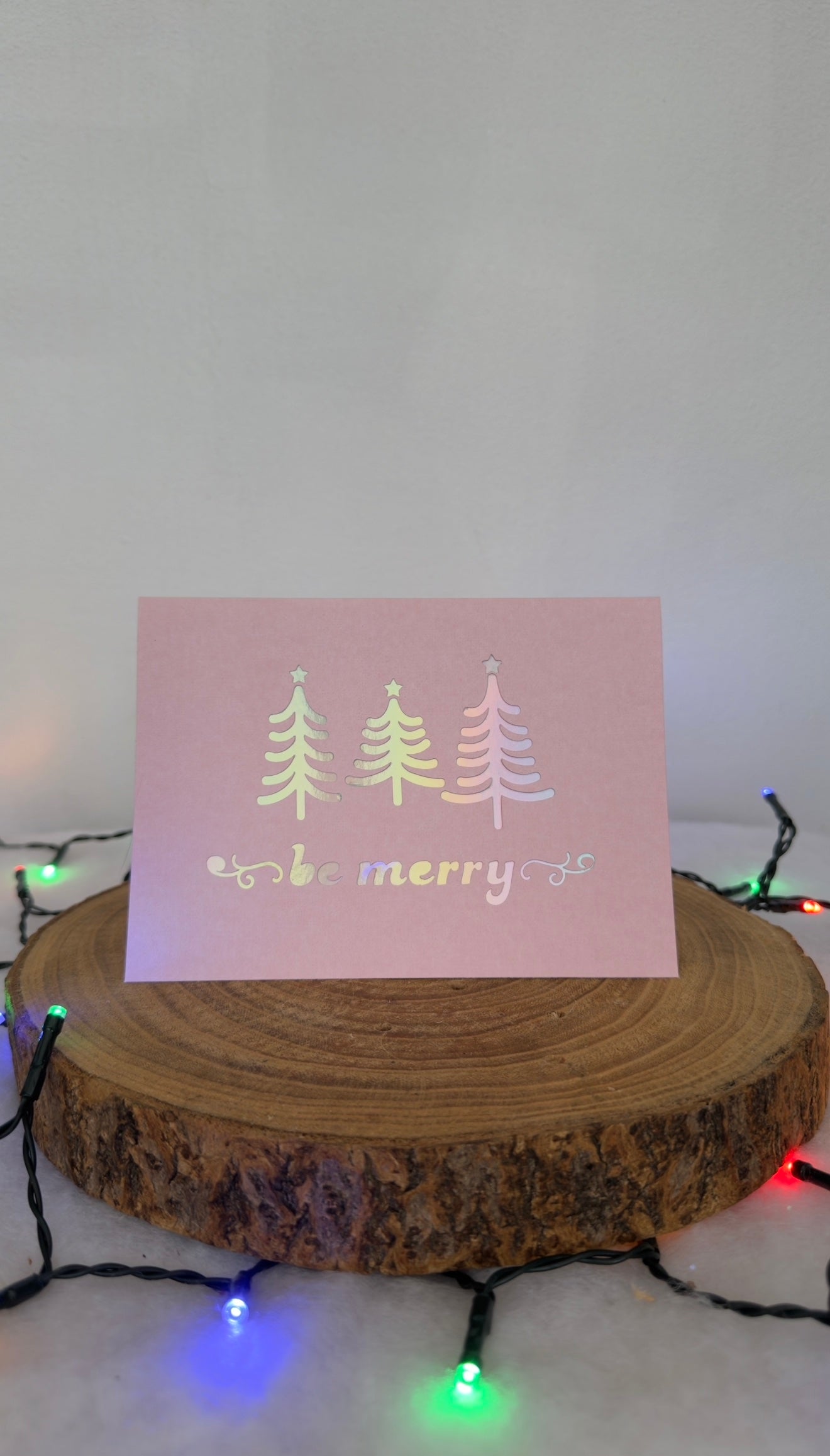 Card with Christmas trees and 'Be Merry' text on a wooden stand with string lights in the background.