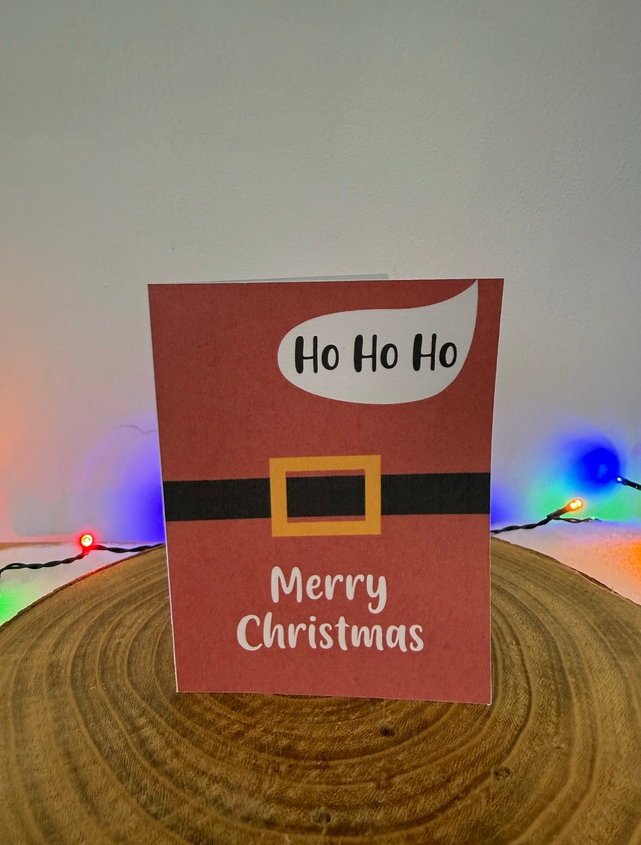 Christmas card with 'Ho Ho Ho' and 'Merry Christmas' text on a red background, placed on a wooden surface with colorful lights in the background.