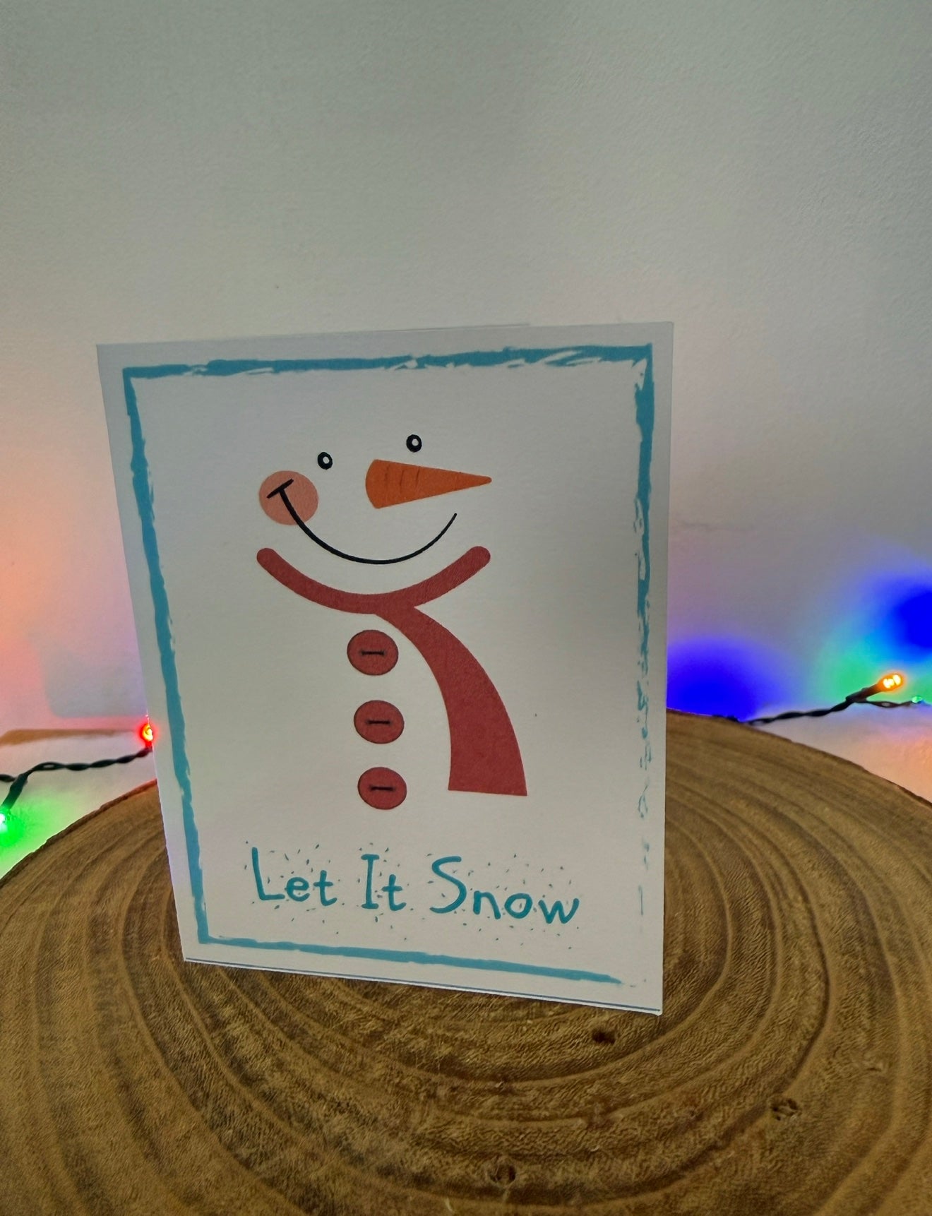 Card with a snowman design and 'Let It Snow' text on a wooden surface with Christmas lights.