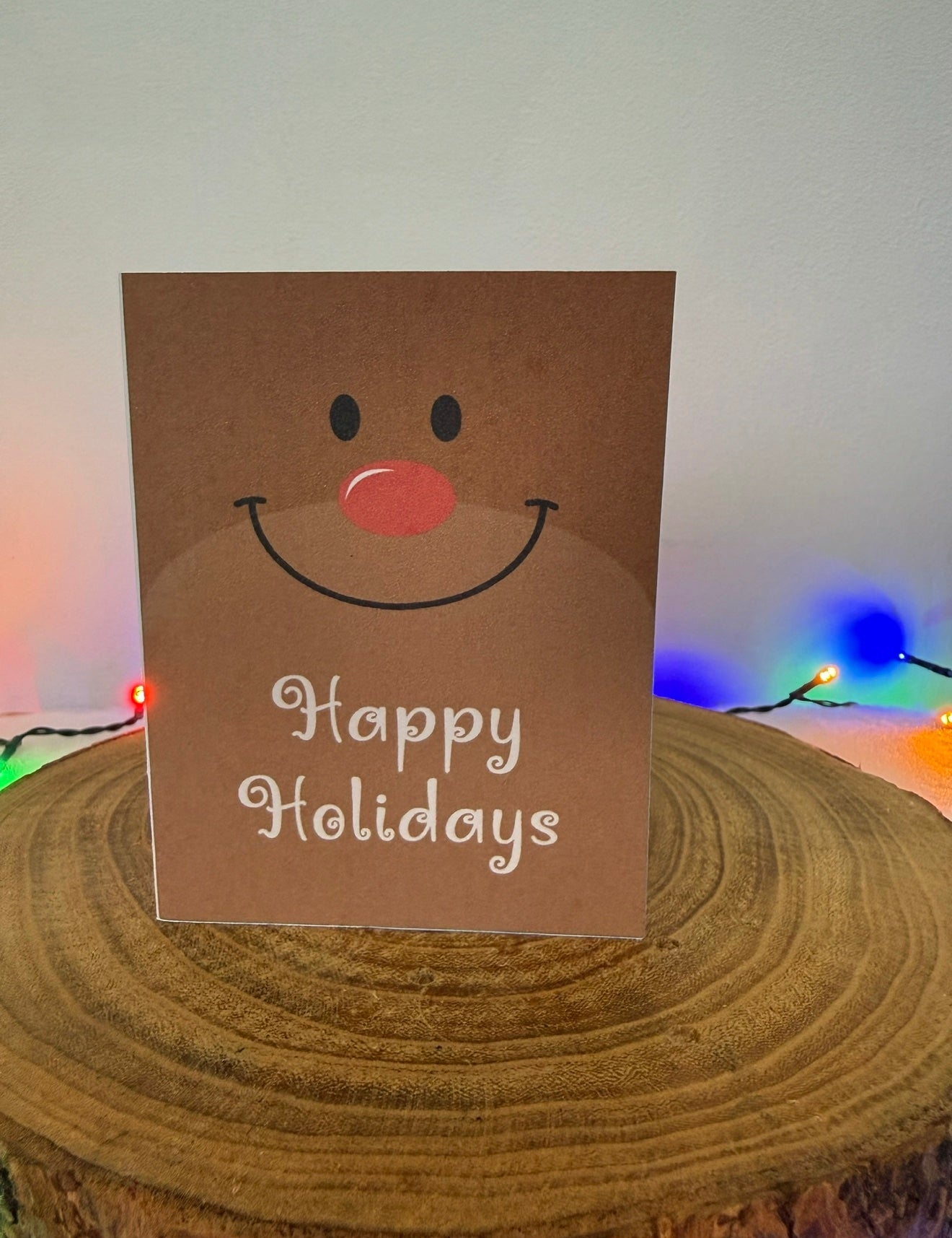Brown greeting card with a face and 'Happy Holidays' text on a wooden surface with string lights in the background.