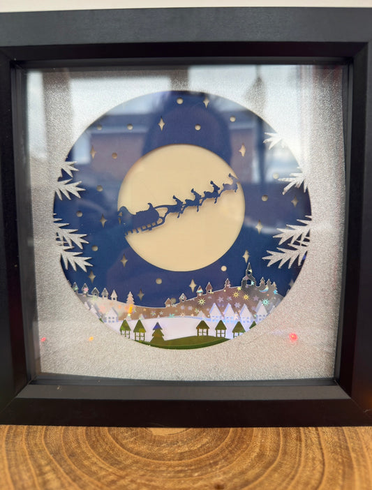 Framed artwork of Santa Claus in a sleigh against a moonlit sky with a town below.