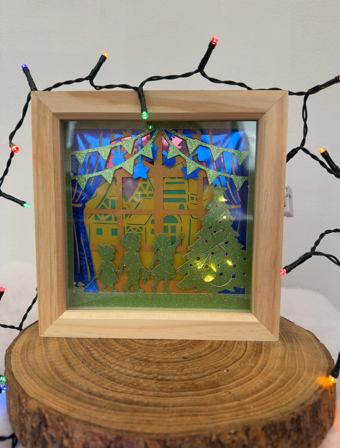 Framed artwork with a house and trees on a wooden stump, surrounded by string lights.