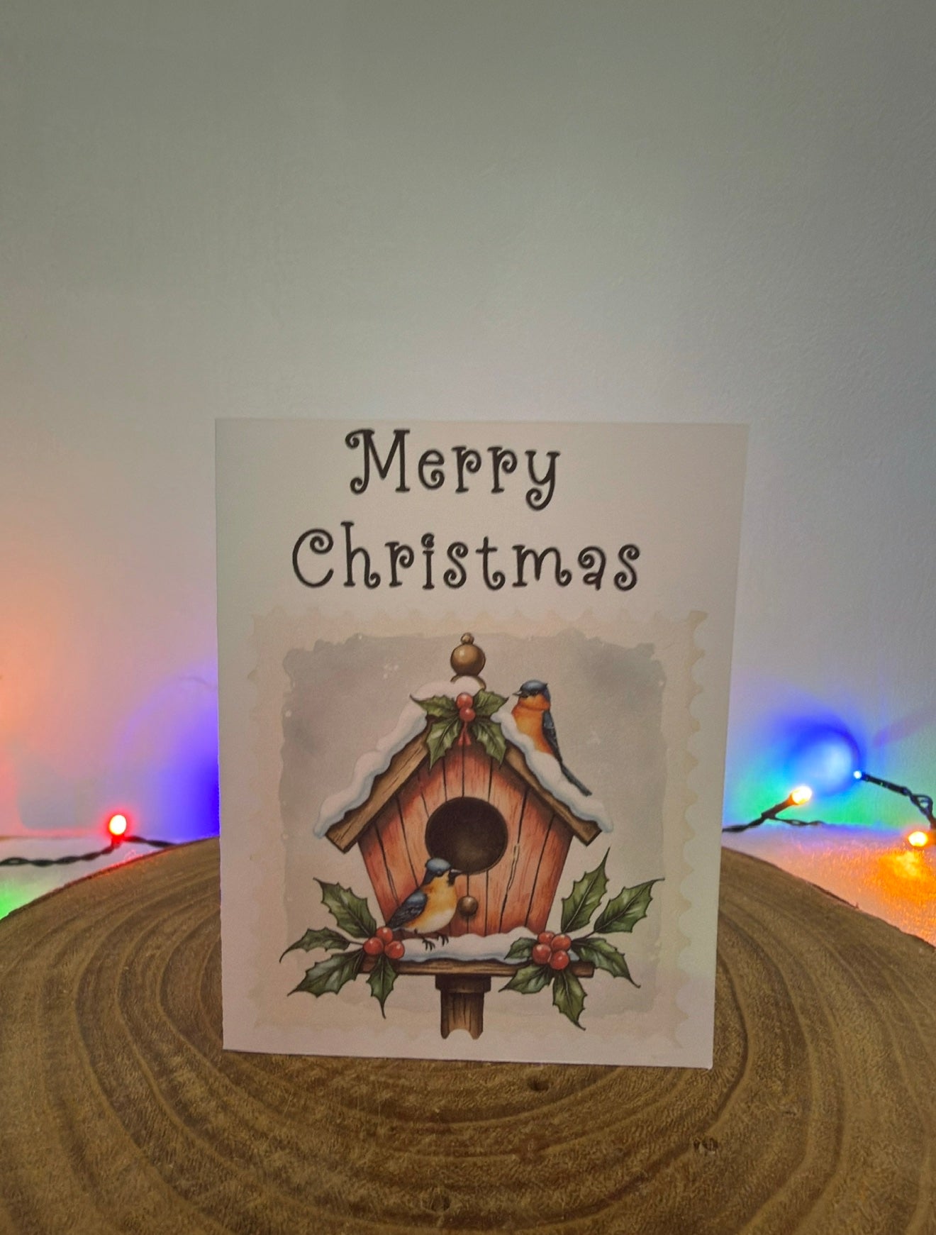 Christmas card with a birdhouse and birds on a wooden surface with festive lights in the background