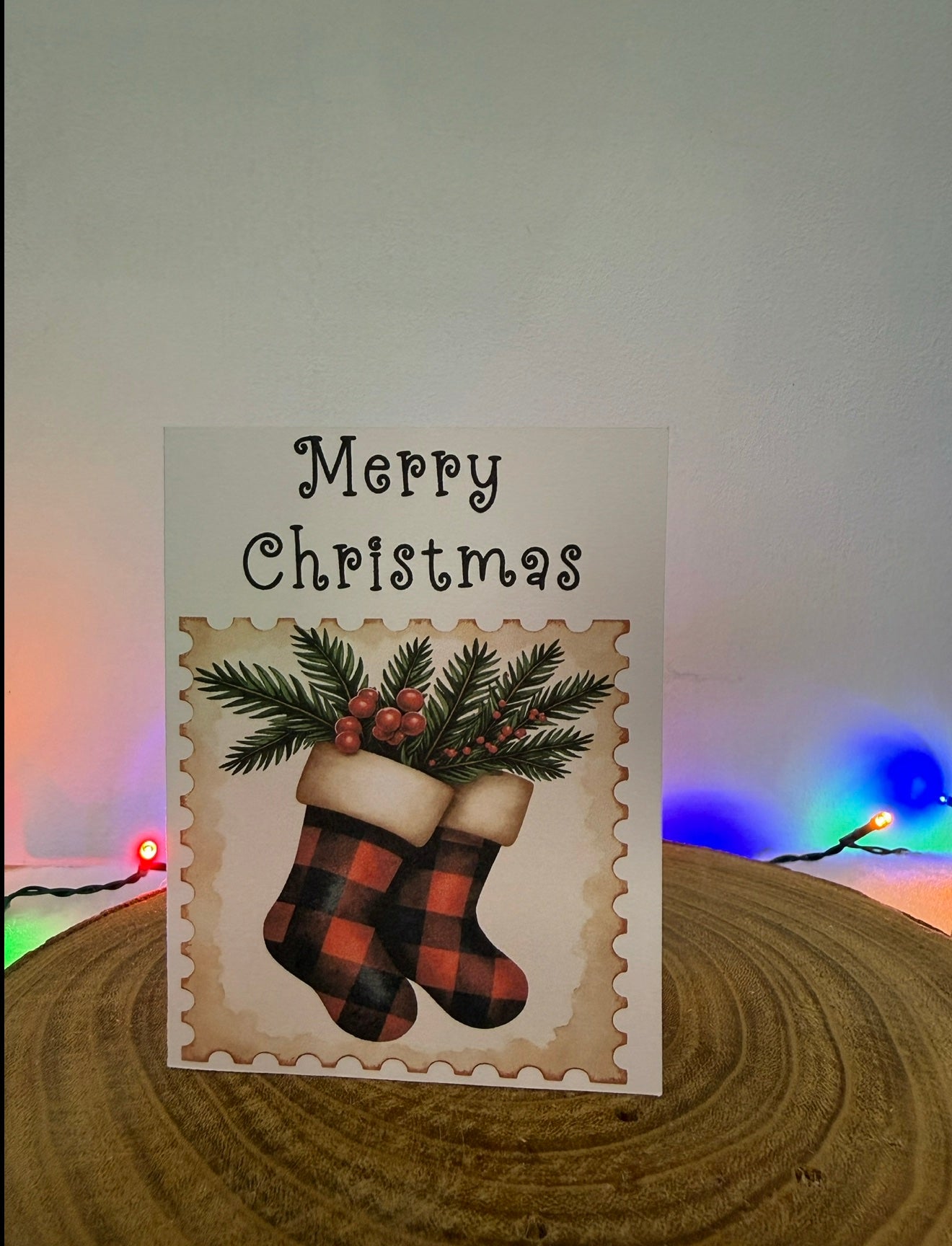 Christmas card with plaid stockings and 'Merry Christmas' text on a wooden surface with festive lights in the background.
