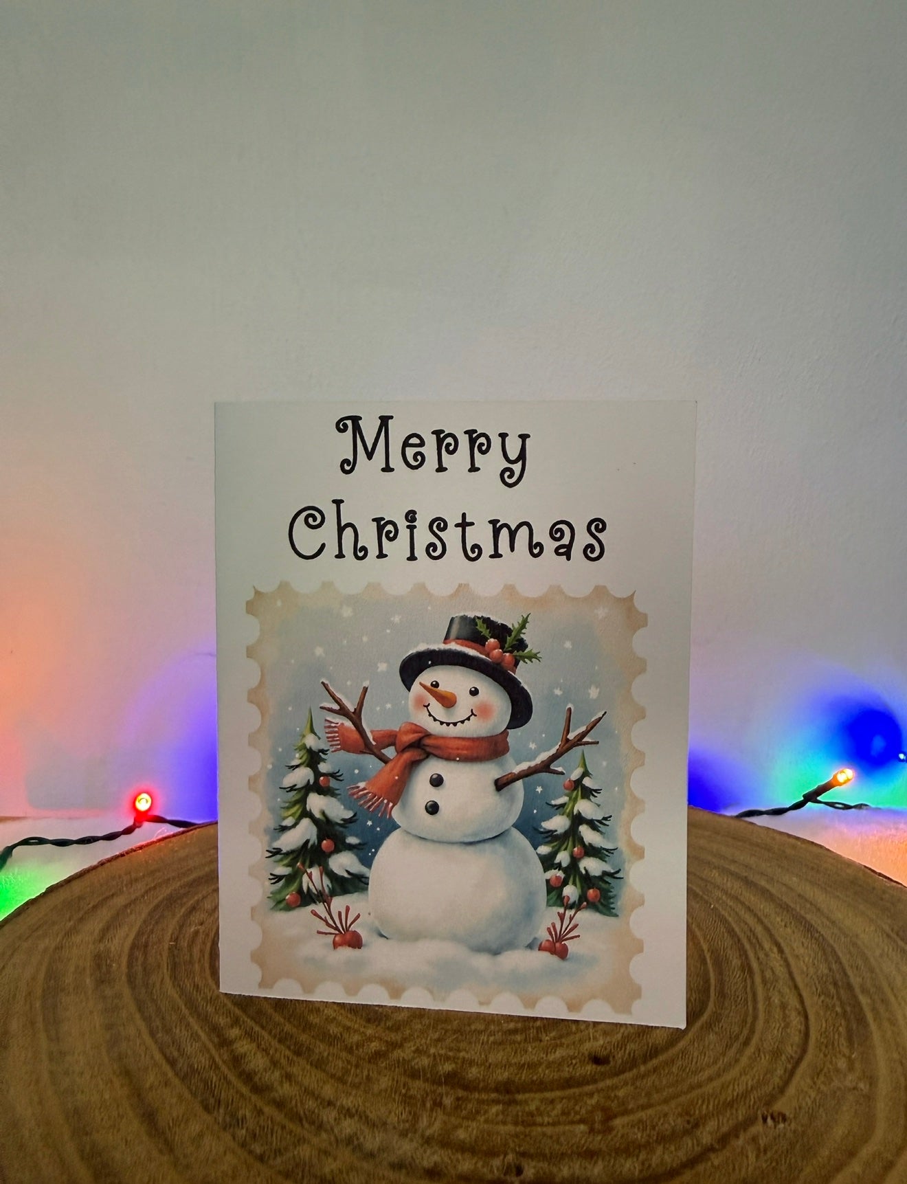 Christmas card with a snowman and 'Merry Christmas' text, placed on a wooden surface with colorful lights in the background.