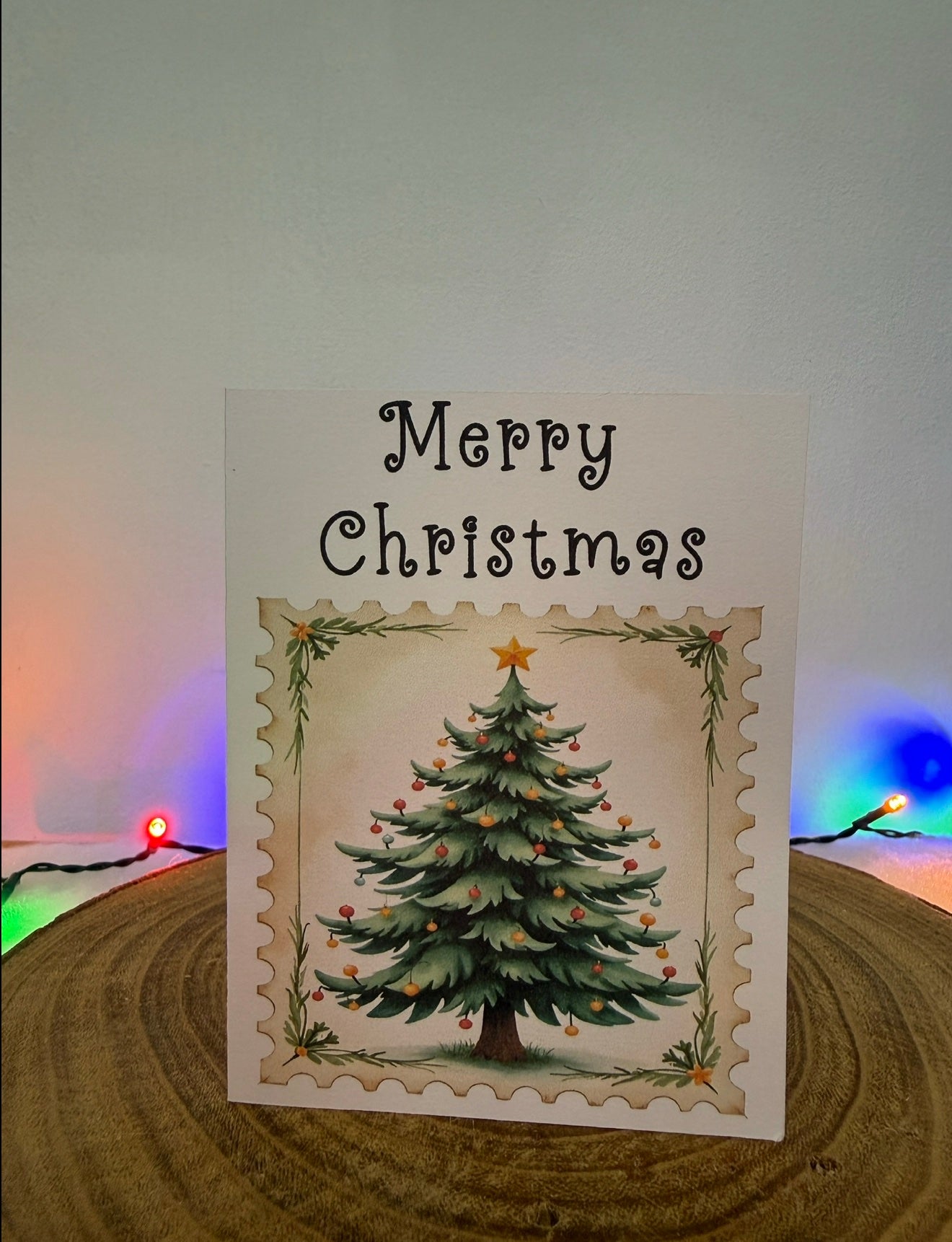 Christmas card with a decorated tree and 'Merry Christmas' text on a wooden surface with colorful lights in the background.