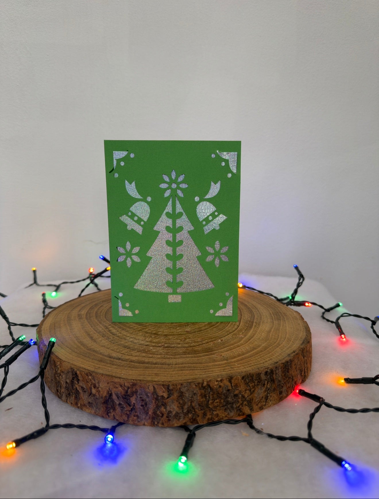Green card with white tree design on a wooden stand with Christmas lights in the background