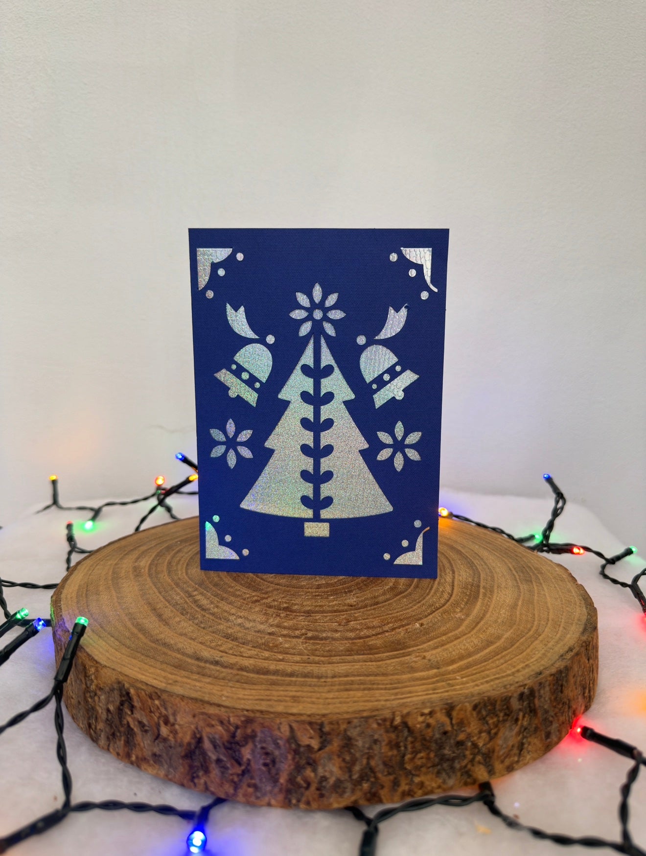 Blue card with white tree design on a wooden stand with Christmas lights in the background