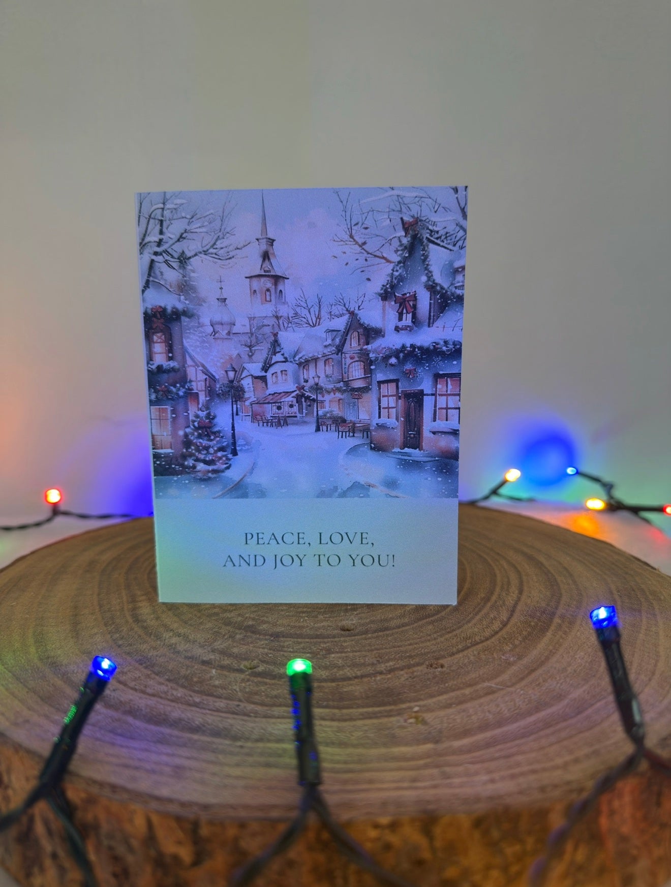 Christmas card with snowy village scene and 'Peace, Love, and Joy to You!' text, placed on a wooden surface with string lights.