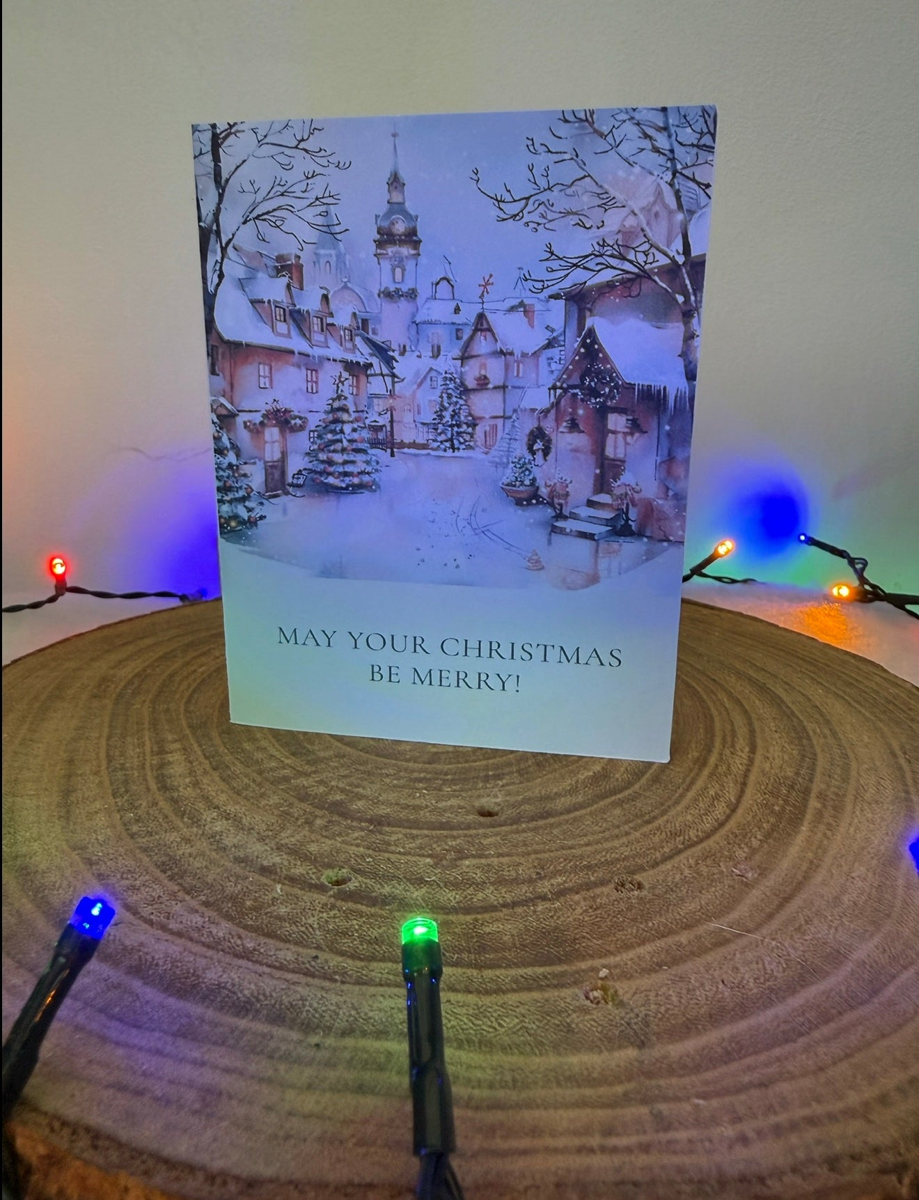 Christmas card with snowy village scene and 'May your Christmas be merry!' text, placed on a wooden surface with festive lights.