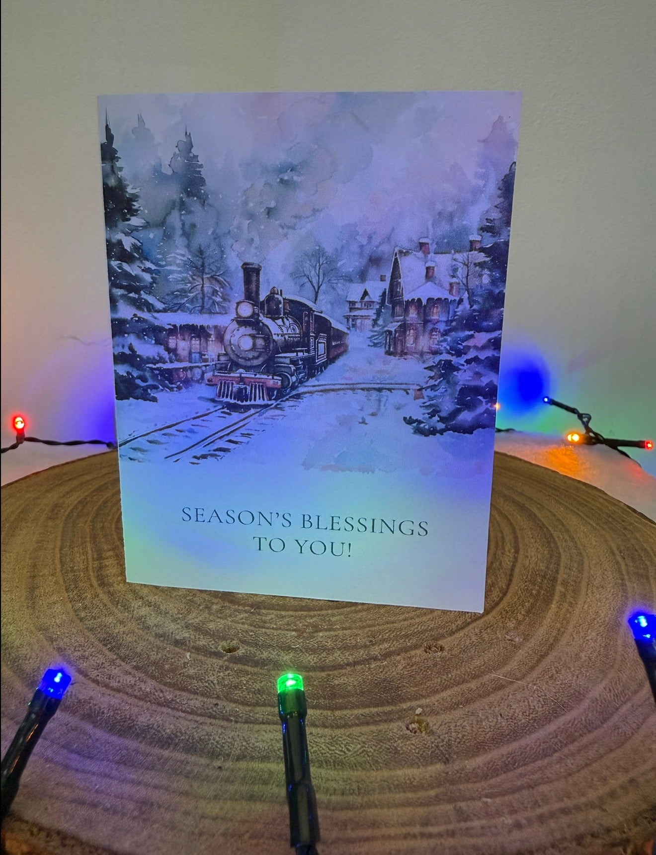 Christmas card with a snowy village and train, surrounded by festive lights on a wooden surface.