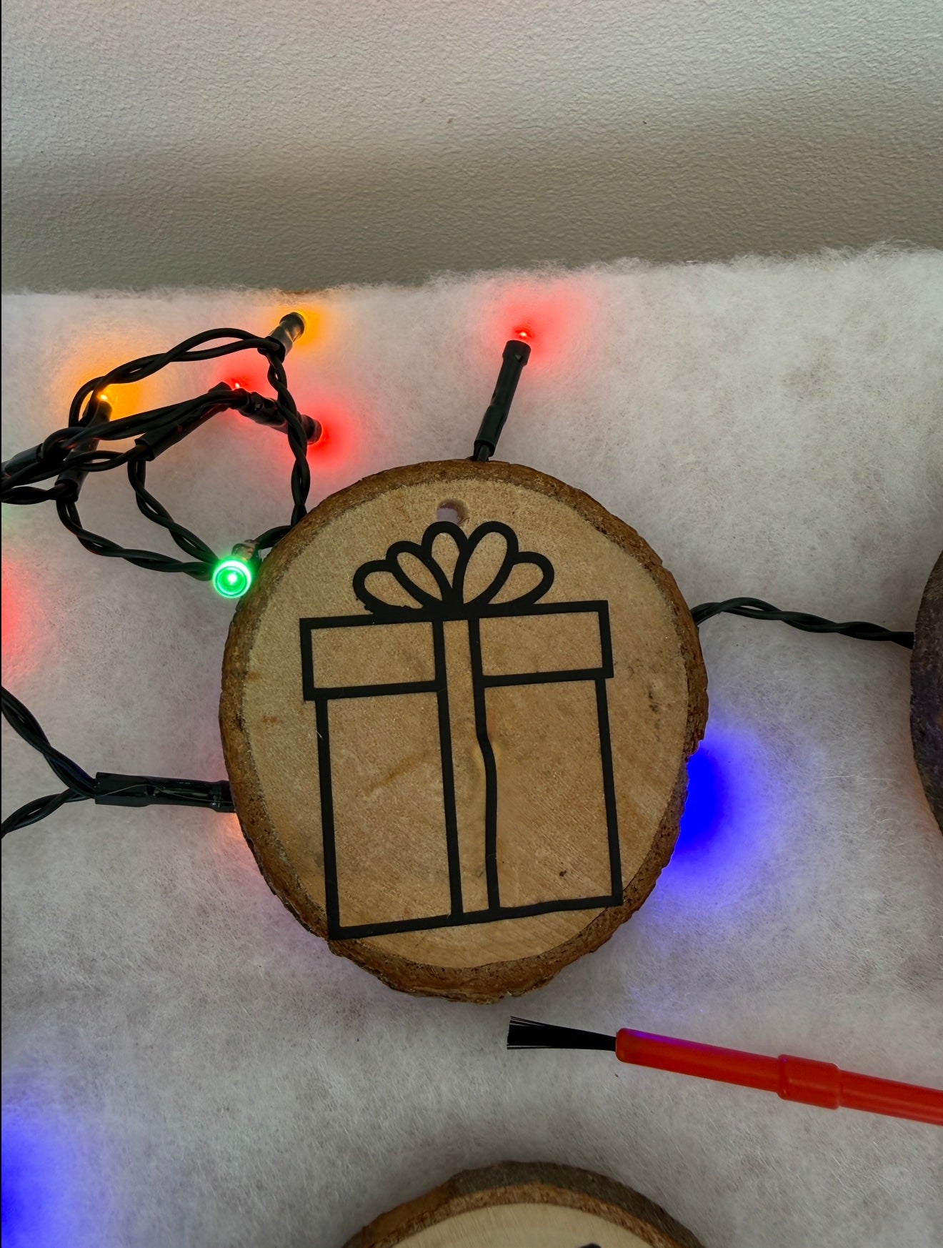 Wooden ornament with a gift box design on a snowy background with Christmas lights.