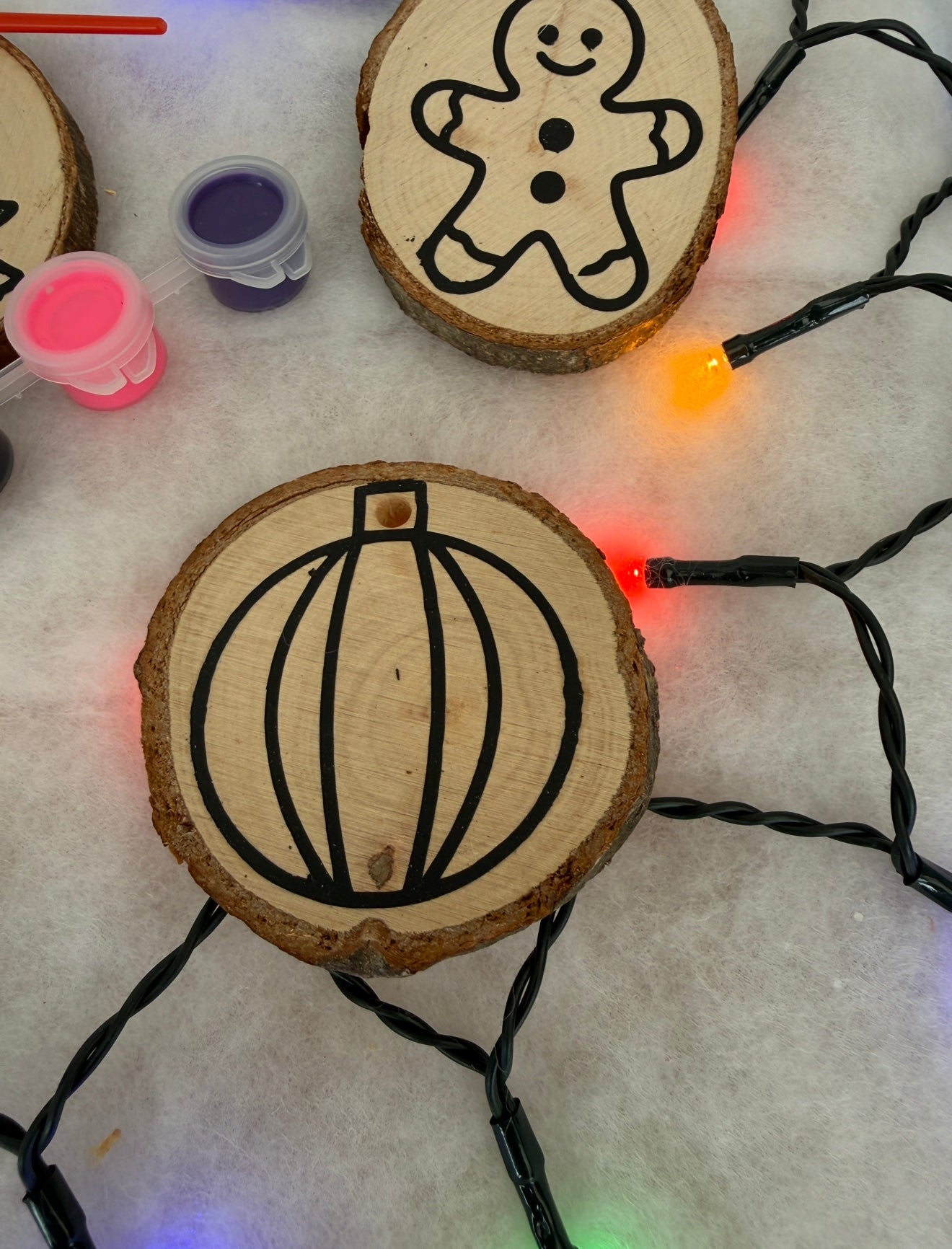 Wooden slices with gingerbread man and pumpkin designs, surrounded by Christmas lights and paint containers.