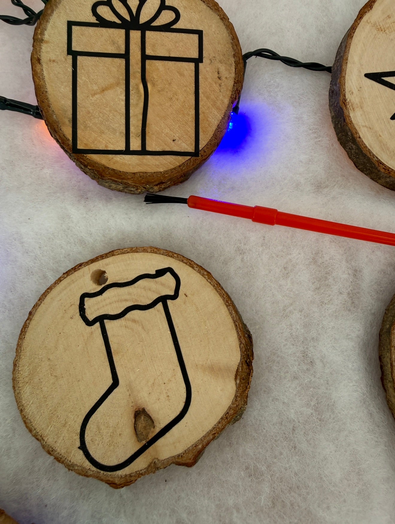 Wooden ornaments with black line drawings of a gift box and a stocking, on a white surface with string lights.