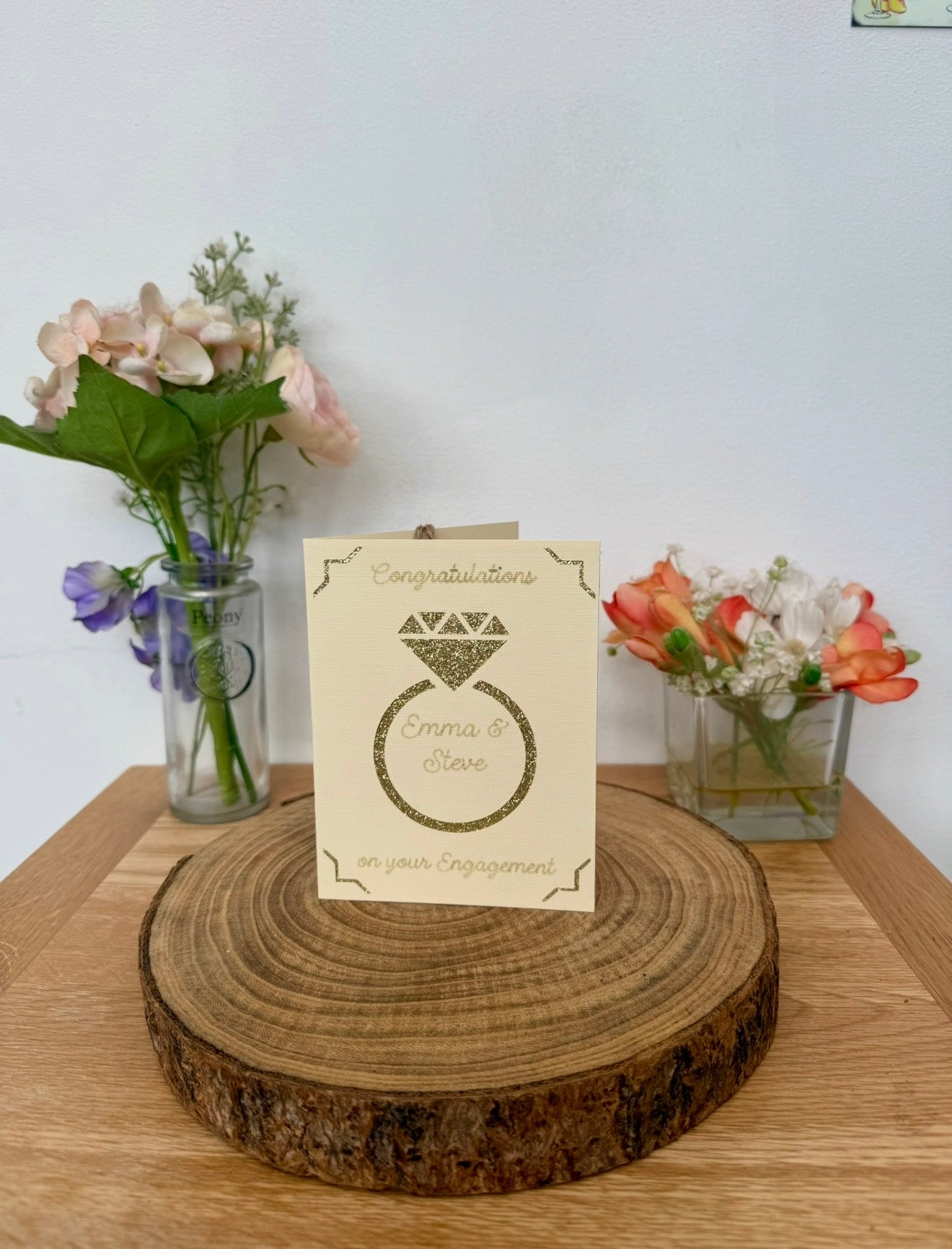 Personalised engagement card with glitter cut-out engagement ring design on cream card. The ring displays the names Emma & Steve inside. Card shown upright on a wooden base with flowers in the background.