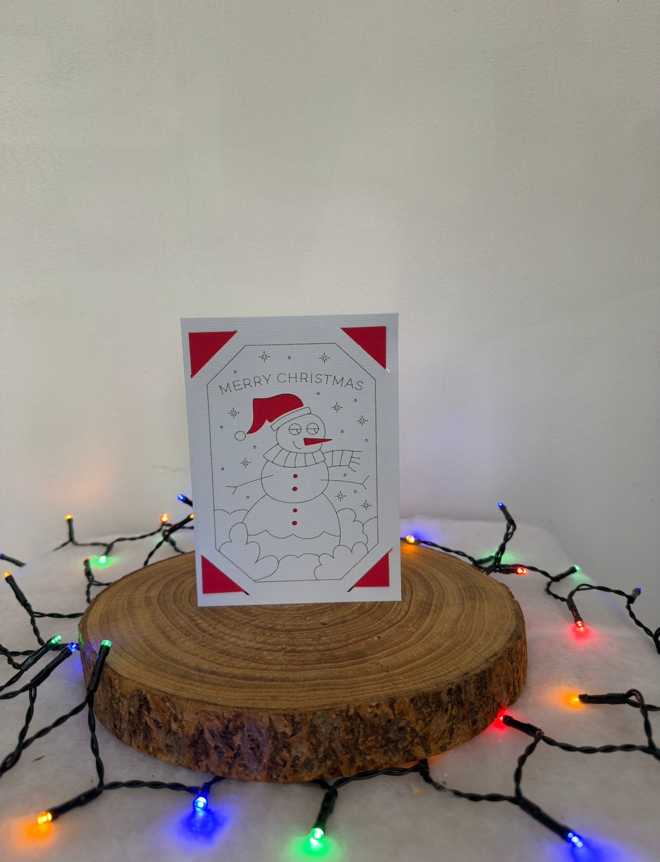 Christmas card with a snowman design on a wooden stand surrounded by festive lights.