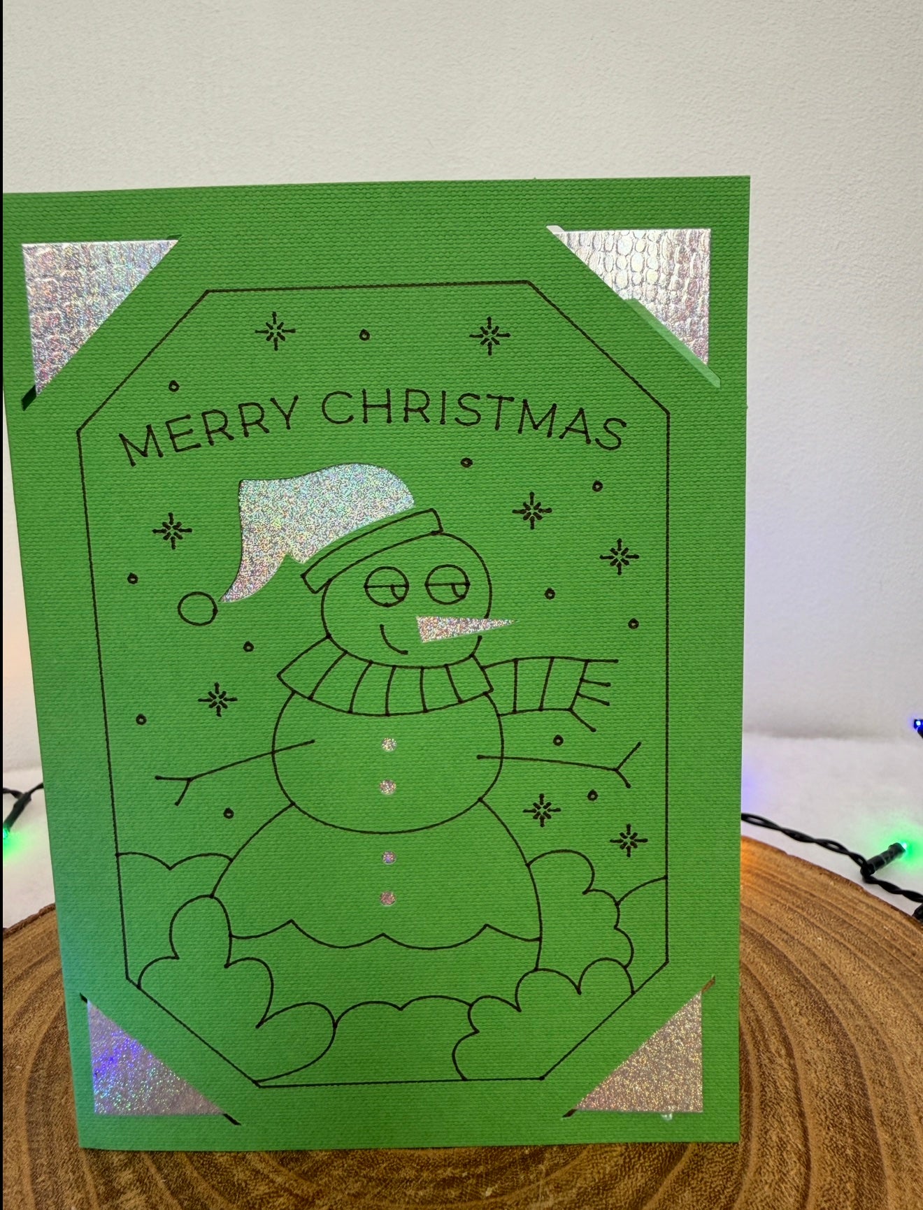 Green Christmas card with a snowman illustration on a wooden surface.