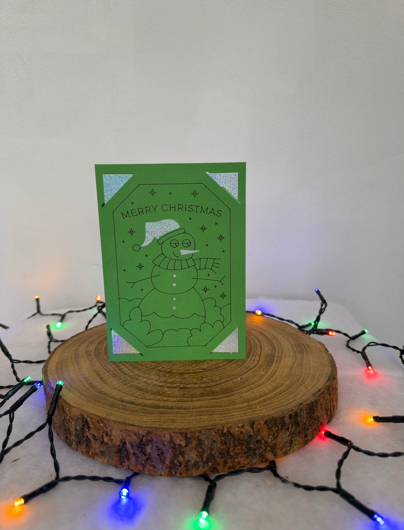 Green Christmas card with a snowman design on a wooden stand with string lights.