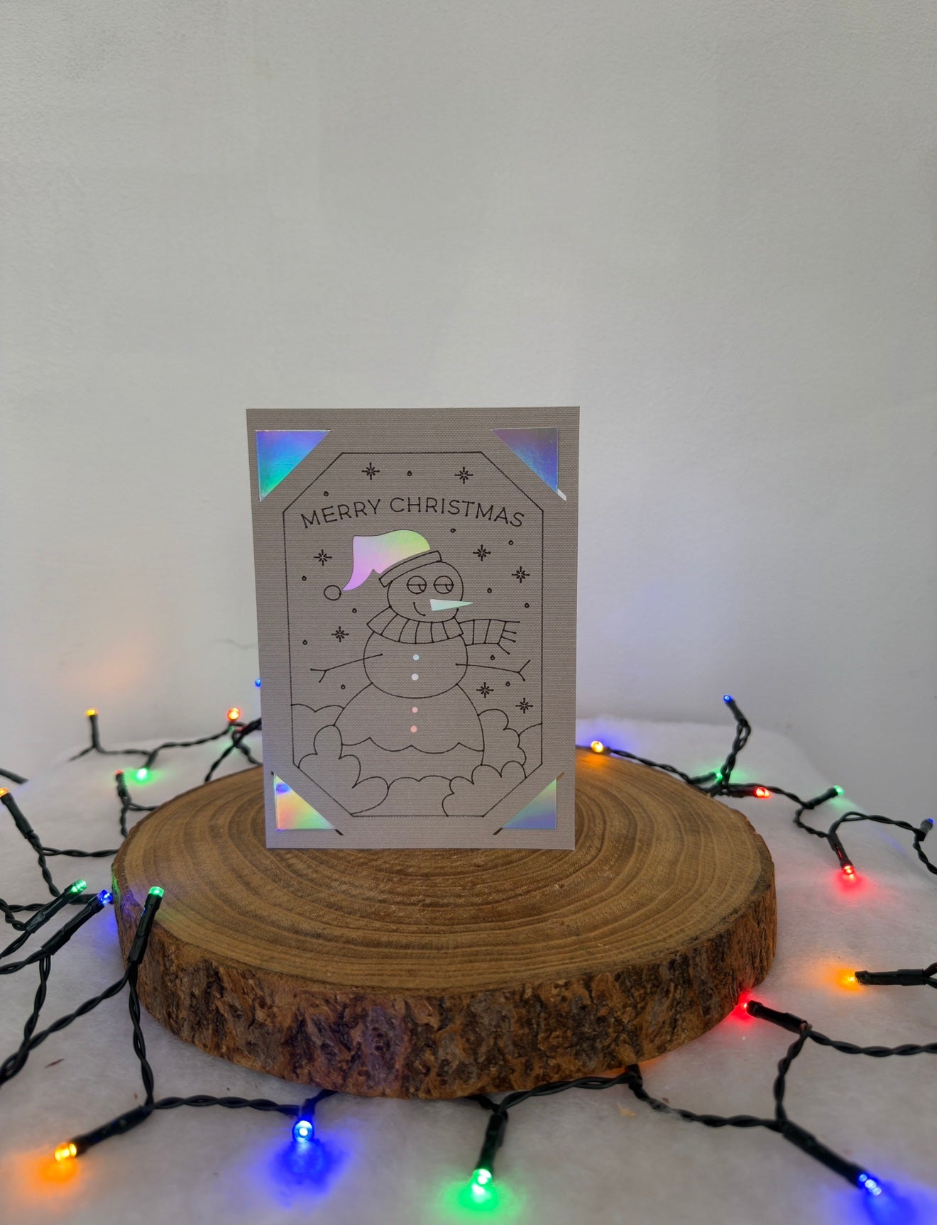 Christmas card with snowman design on a wooden stand surrounded by colorful lights.