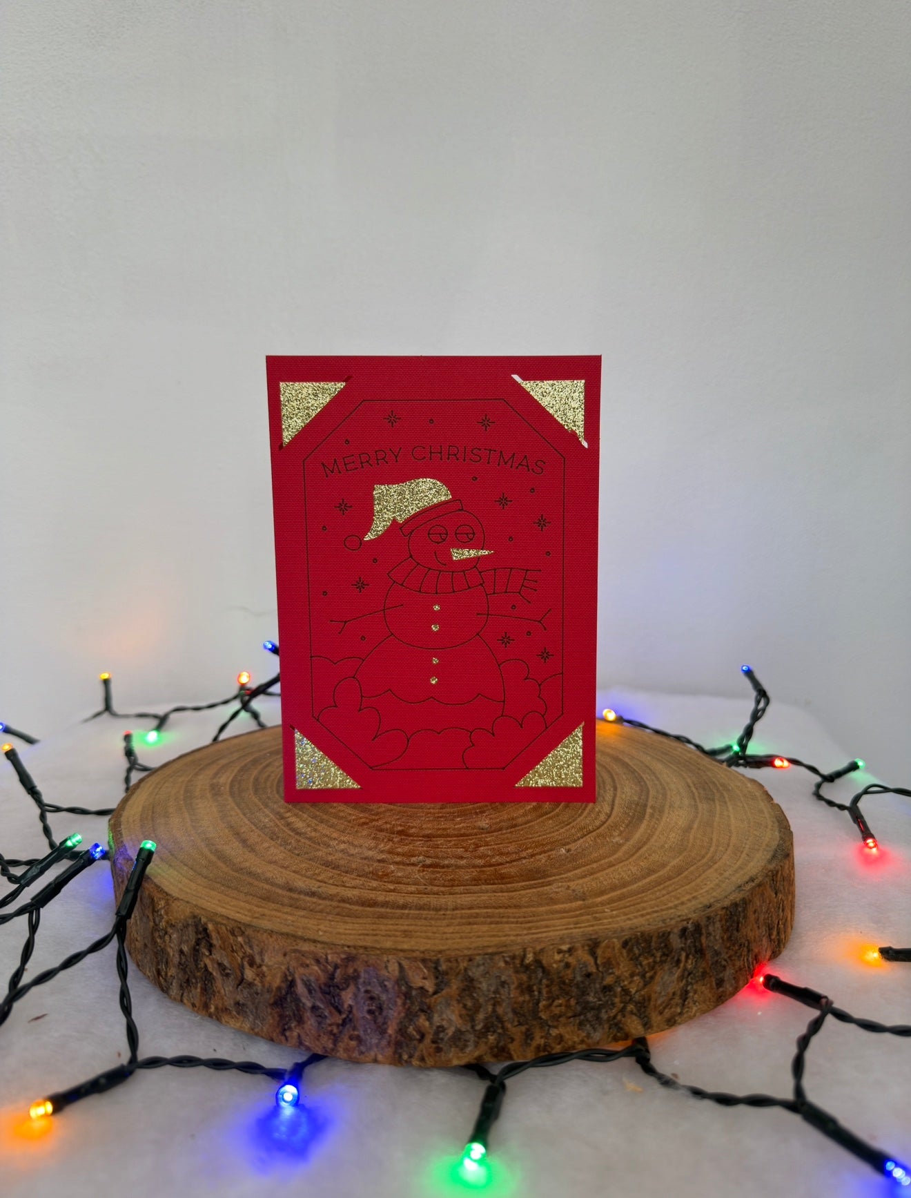 Red Christmas card with gold accents on a wooden stand surrounded by colorful lights.