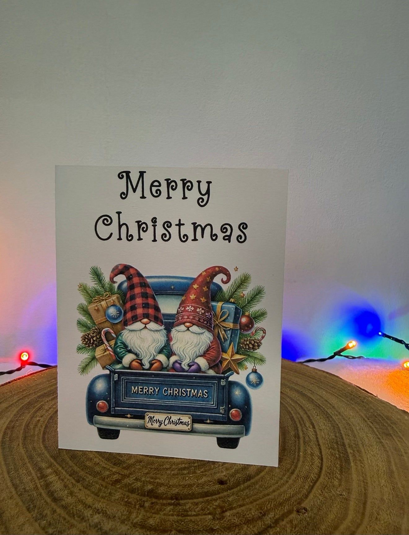 Christmas card with two gnomes in a truck and 'Merry Christmas' text, placed on a wooden surface with festive lights in the background.