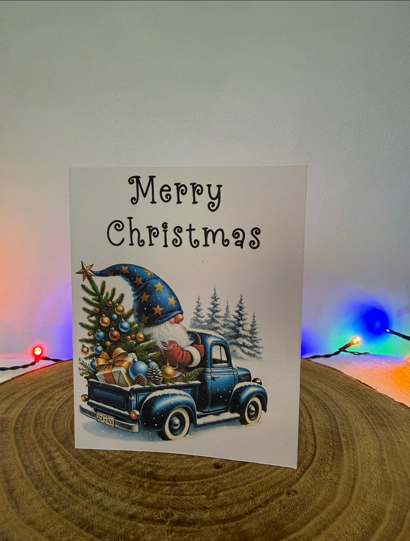 Christmas card with a gnome driving a truck and 'Merry Christmas' text, placed on a wooden surface with blurred lights in the background.