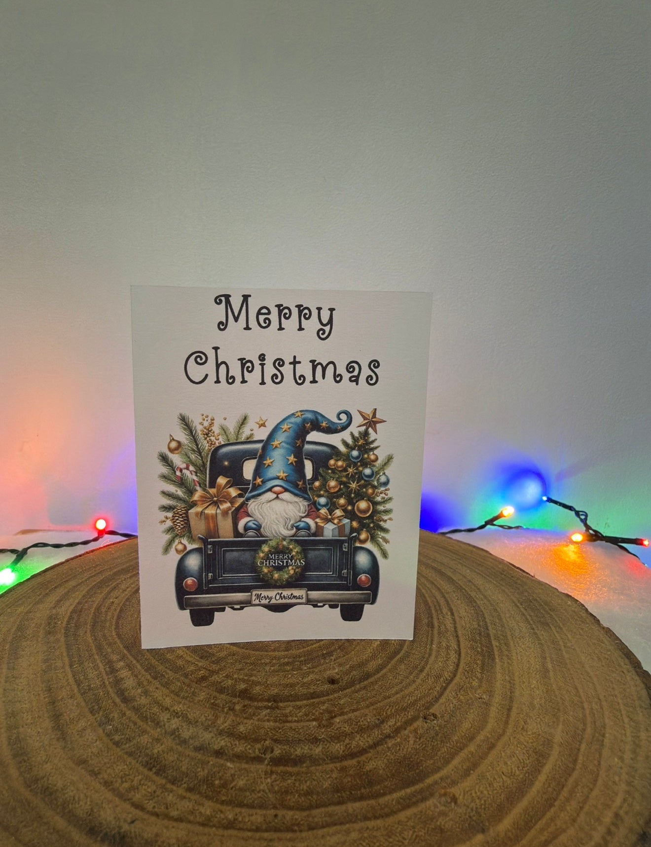 Christmas card with a gnome in a truck and 'Merry Christmas' text, placed on a wooden surface with colorful lights in the background.