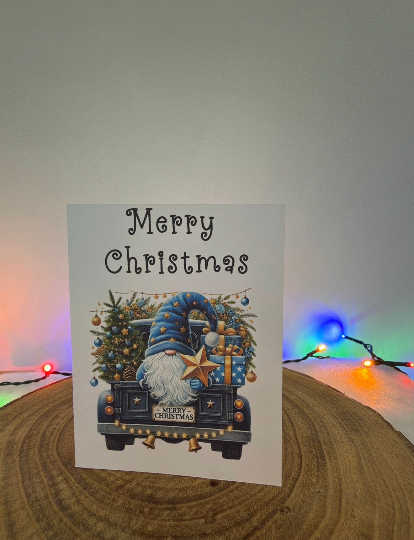 Christmas card with a gnome driving a truck and 'Merry Christmas' text, placed on a wooden surface with colorful lights in the background.