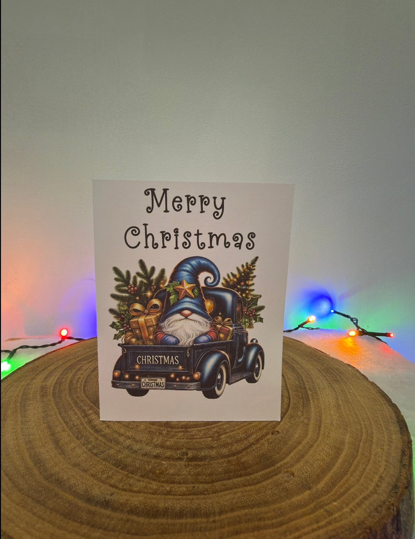 Christmas card with a gnome and car illustration on a wooden surface with colorful lights in the background.