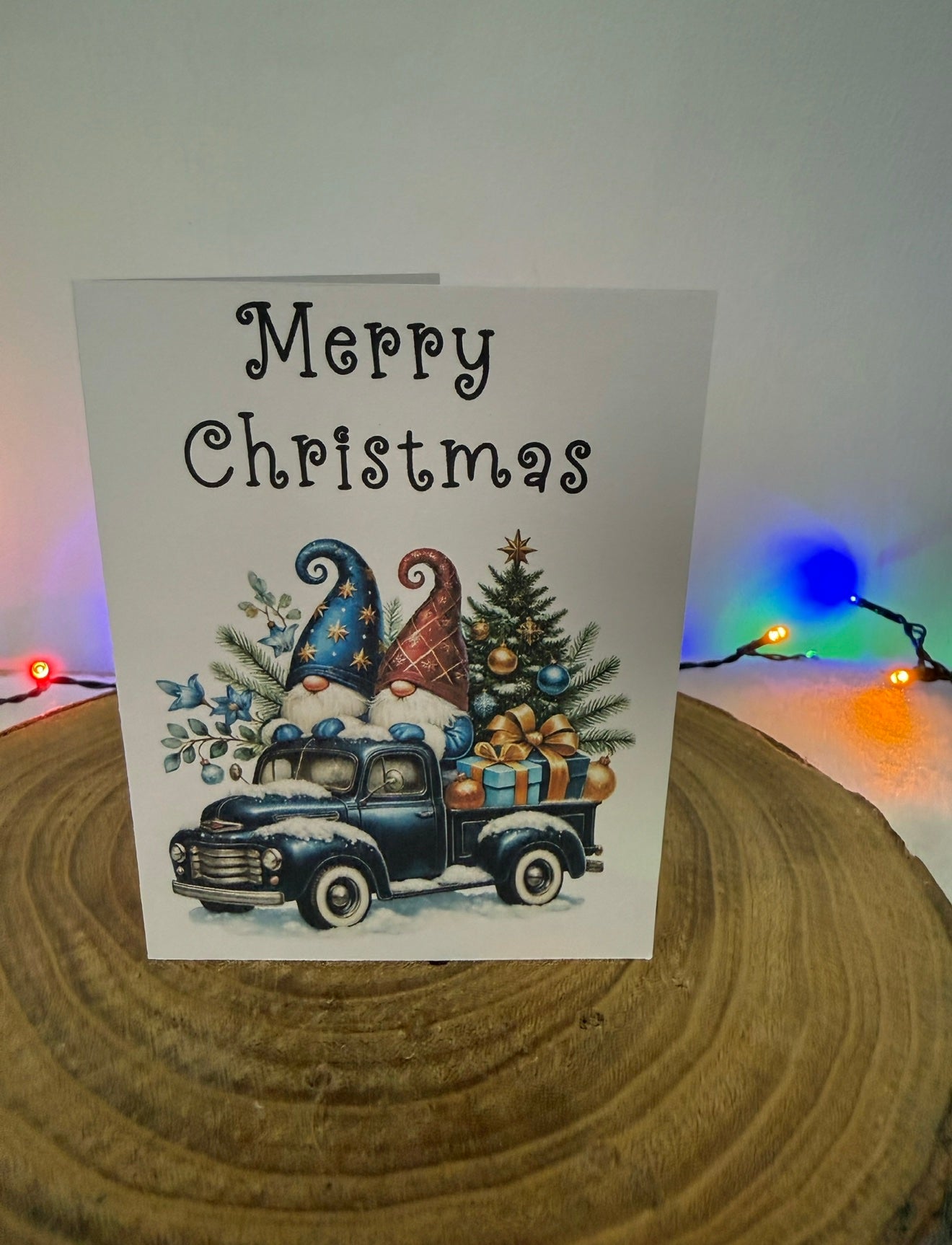 Christmas card with gnomes, truck, and tree on a wooden surface with festive lights in the background.