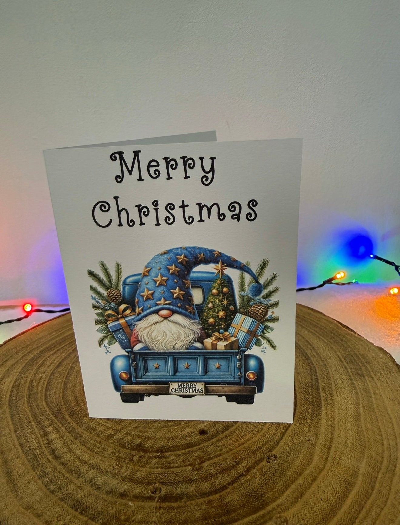 Christmas card with a blue gnome driving a truck and 'Merry Christmas' text, placed on a wooden surface with festive lights in the background.