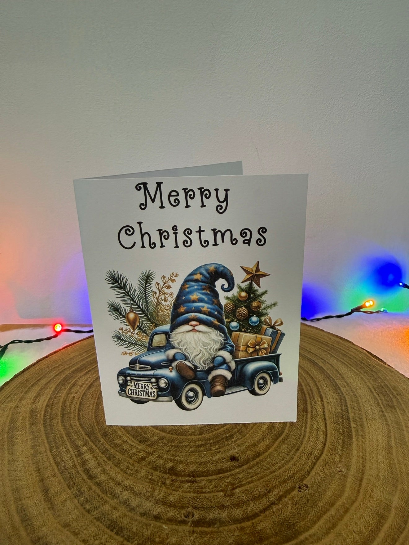 Christmas card with a gnome driving a truck and 'Merry Christmas' text, placed on a wooden surface with colorful lights in the background.