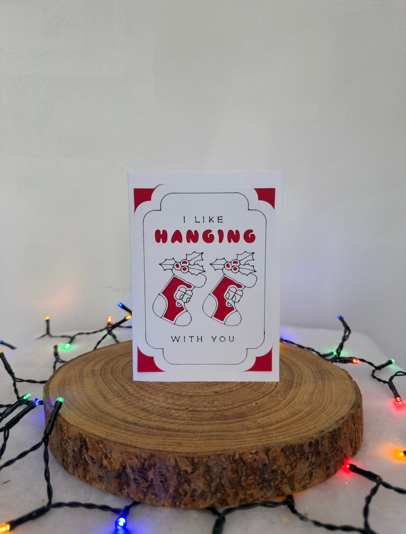 Card with stockings and text 'I like hanging with you' on a wooden stand with Christmas lights in the background.