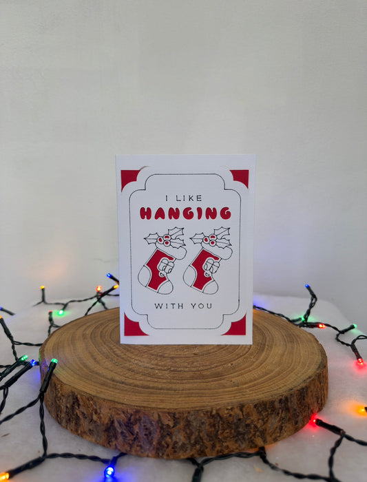 Card with stockings and text 'I like hanging with you' on a wooden stand with Christmas lights in the background.
