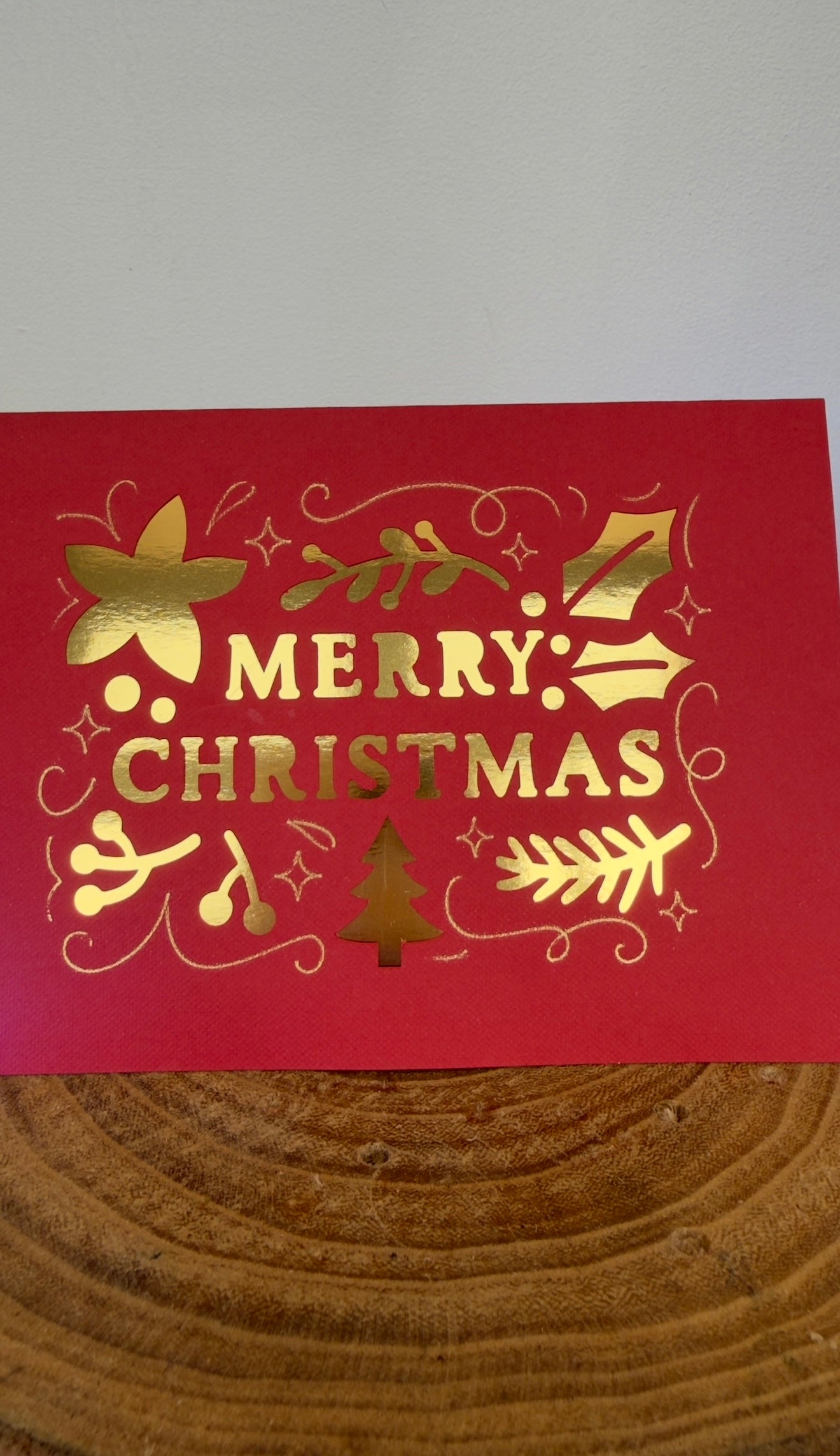 Red Christmas card with gold 'Merry Christmas' text and decorative elements on a wooden surface.