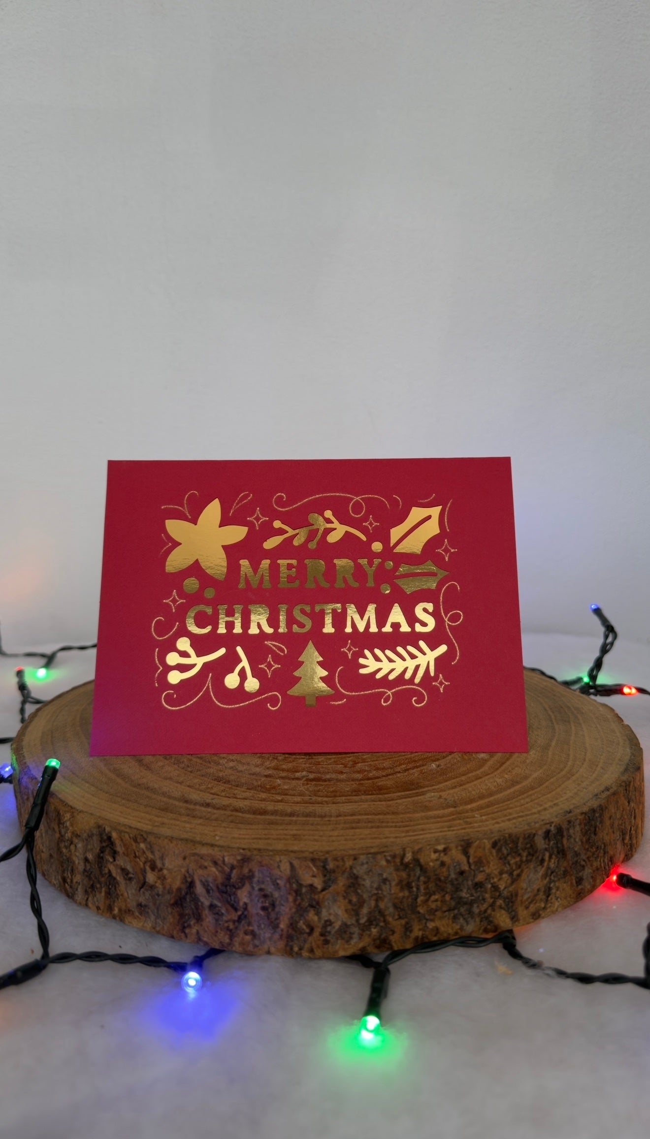 Red Christmas card with gold text and decorations on a wooden stand with festive lights in the background.
