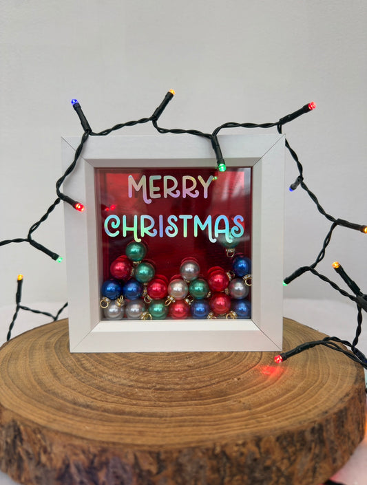 Framed 'Merry Christmas' sign with colorful ornaments and string lights on a wooden surface.
